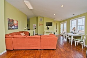 A living room with a sectional sofa, a dining table with chairs, a TV mounted on the wall, and a window with a view. - 1 Atlantic View, Oceanfront Home in Emerald Isle, NC! (Emerald Isle)