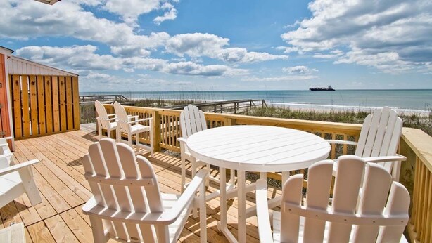 A wooden deck with white Adirondack chairs and a round table, overlooking a beach and ocean. - 2 Seas the Day West, Oceanfront Duplex in Emerald Isle, NC! (Emerald Isle)