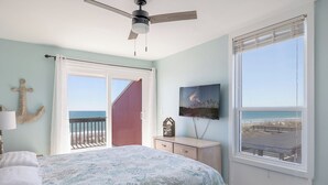 A bedroom with a bed, a ceiling fan, a TV, and a large window with a view of the beach. - 2 Seas the Day West, Oceanfront Duplex in Emerald Isle, NC! (Emerald Isle)