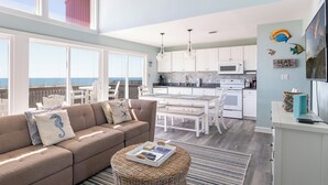 A modern living room with a large window overlooking the sea, a kitchen with white cabinets, and a dining area with white furniture. - 2 Seas the Day West, Oceanfront Duplex in Emerald Isle, NC! (Emerald Isle)