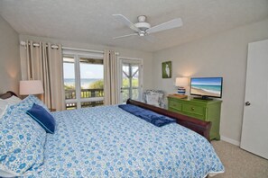 A bedroom with a large bed, a ceiling fan, a television, and a view of the ocean through the window.