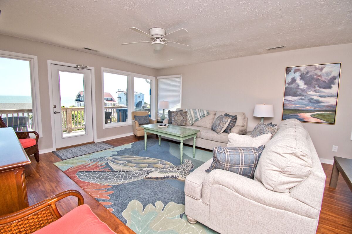 A spacious living room with a view of the ocean, a ceiling fan, a sofa, a coffee table, and a rug with a leaf pattern.