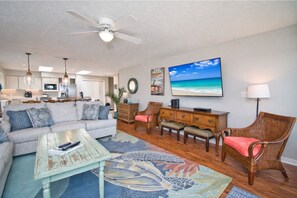 A spacious living room with a sofa, coffee table, and wicker chairs. A television and artwork are on the wall.