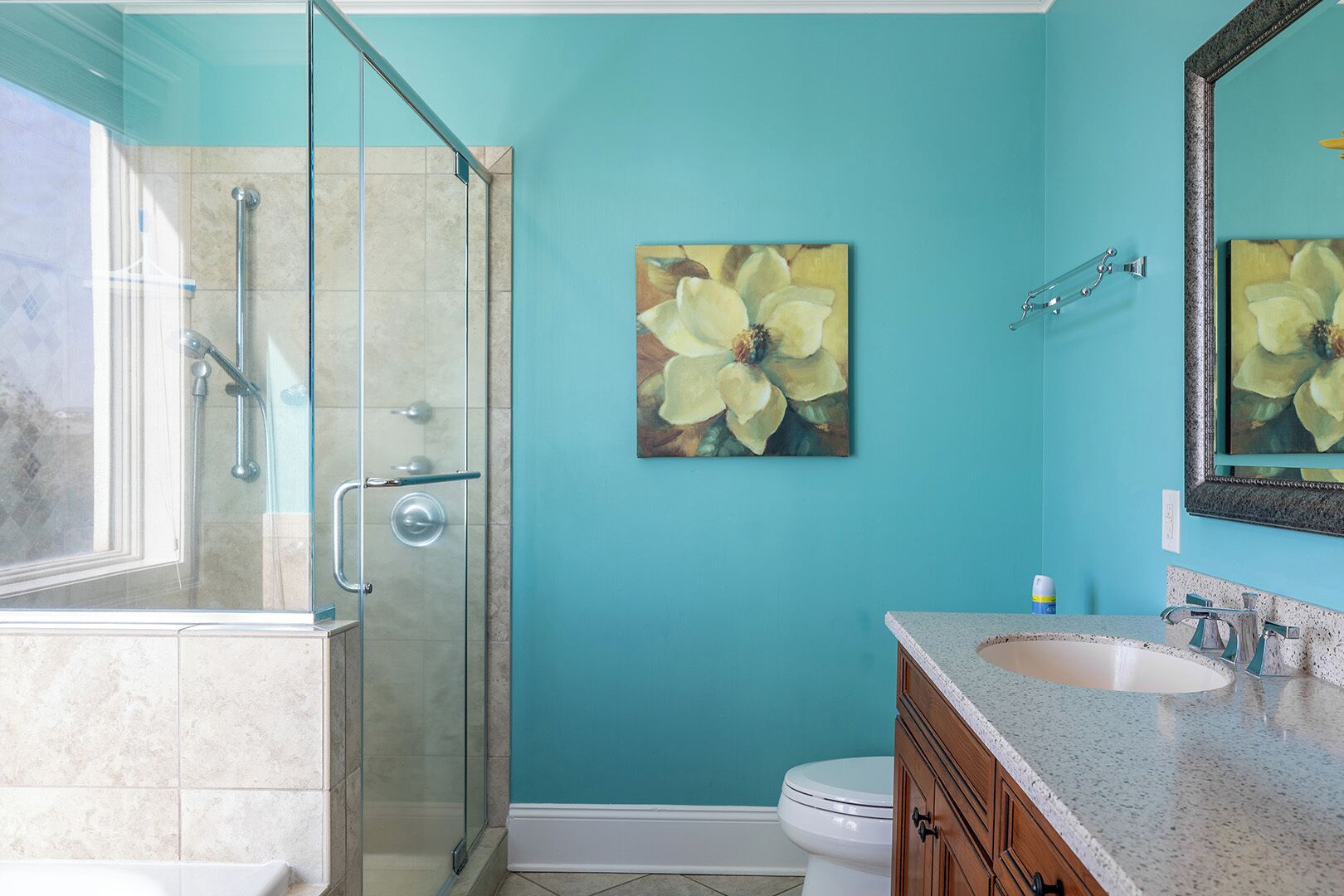 A bathroom with a glass shower enclosure, a white toilet, a wooden vanity with a granite countertop, and a large floral painting on the wall.