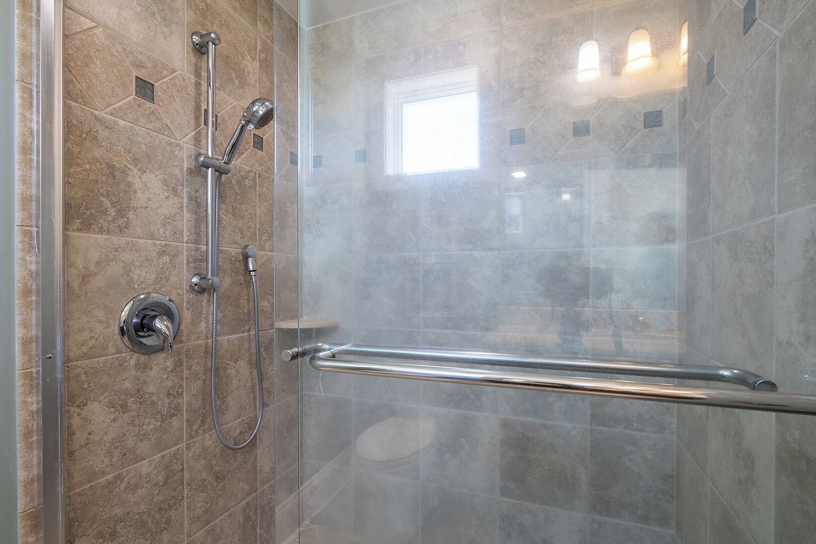 Une douche fermée par des parois en verre, dotée d’un pommeau de douche à main et d’un flexible de douche à main, ainsi que d’une barre d’appui et d’une petite fenêtre.