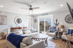 A beach house interior with a sofa, armchairs, a coffee table, and a view through the glass door. - A Pirates Dream, Almost Oceanfront House in Emerald Isle, NC! (Emerald Isle)