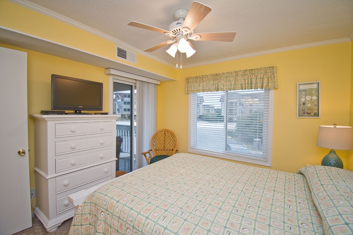 A bedroom with a bed, a dresser, a television, a ceiling fan, and a window with blinds.