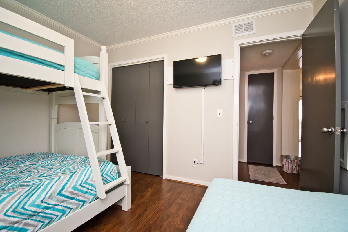 A bedroom with a bunk bed, a single bed, a TV mounted on the wall, and a door leading to another room.