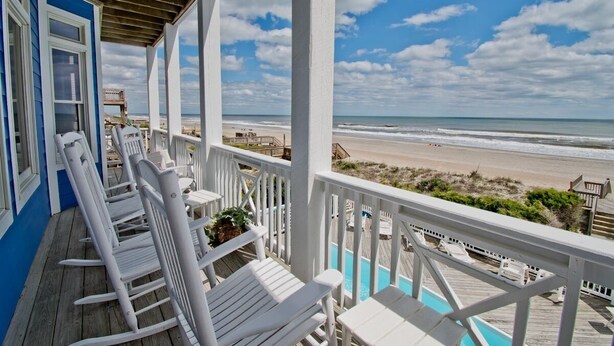 Une terrasse en bois avec des chaises blanches et une piscine, donnant sur une plage.