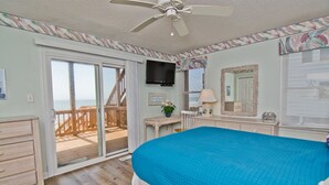 A bedroom with a large bed, a ceiling fan, a television, and a balcony with a wooden deck. - Agape West, Oceanfront Duplex in Emerald Isle, NC! (Emerald Isle)