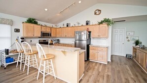 A modern kitchen with wooden cabinets, a central island with bar stools, stainless steel appliances, and decorative plants. - Agape West, Oceanfront Duplex in Emerald Isle, NC! (Emerald Isle)
