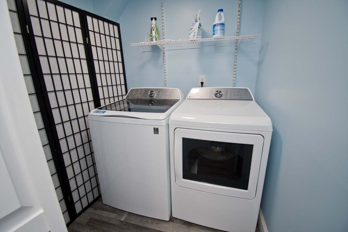 A laundry room with two washing machines and a shelf above them holding bottles.