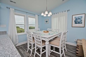 Un coin repas avec une table et des chaises blanches, un lustre et une vue sur la verdure à l’extérieur.