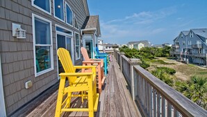 Una terraza de madera con sillas Adirondack de colores, barandilla de madera y vista a casas y follaje.