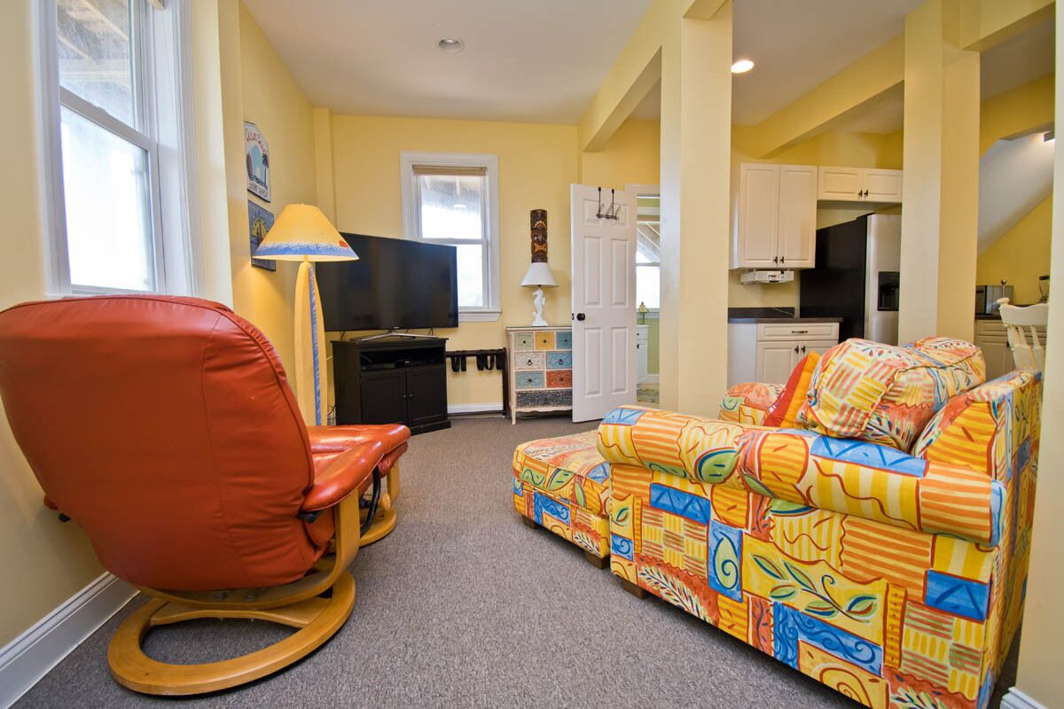 A living room with a red rocking chair, a colorful sofa, a television, and a kitchen area in the background.