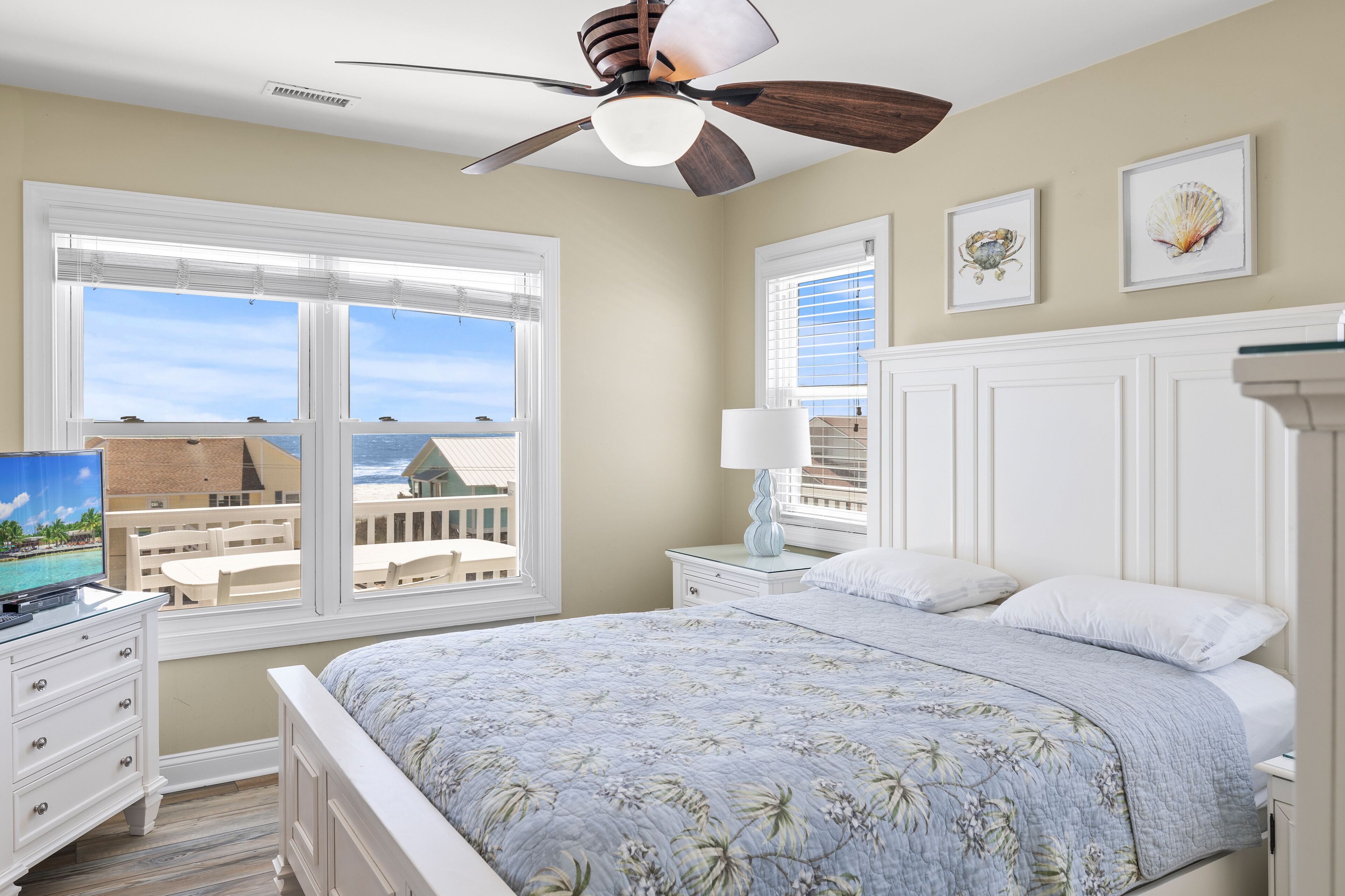 A bedroom with a bed, a ceiling fan, a window with a view of buildings, a nightstand with a lamp, and a dresser.