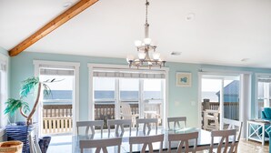 A dining area with a glass table and chairs, large windows with a view of the ocean, and a wooden ceiling. - Angel's Dream, Oceanfront House in Emerald Isle, NC! (Emerald Isle)