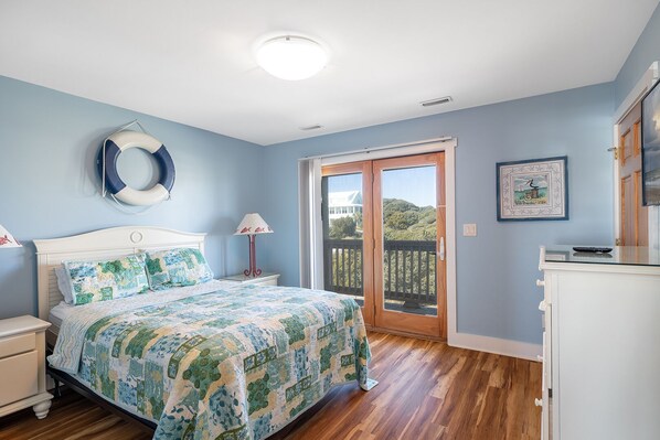 A bedroom with a bed, a nightstand, a dresser, a window with a view of a house and trees, and a door to an outdoor balcony. - Angel's Dream, Oceanfront House in Emerald Isle, NC! (Emerald Isle)