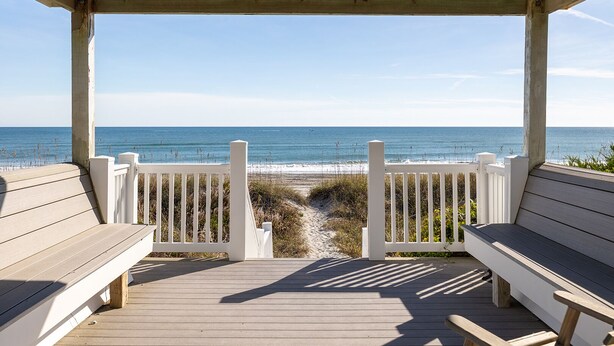 Une terrasse en bois avec des rampes blanches qui mène à une plage, à des bancs et offre une vue dégagée sur l’océan.