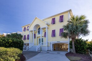 A large, yellow building with purple shutters, a central entrance with steps, and a palm tree in the foreground. - Angel's Haven, Oceanfront House in Emerald Isle, NC! (Emerald Isle)