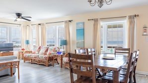 A spacious living room with a wooden dining table, wicker furniture, and large windows with curtains. - Anniversary Waltz East, Oceanfront Duplex in Emerald Isle, NC! (Emerald Isle)