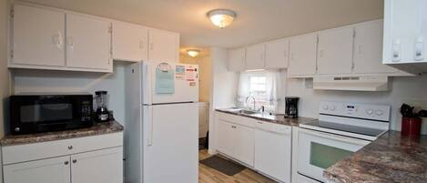 Una cocina compacta con gabinetes blancos, microondas, refrigerador, estufa y cafetera.
