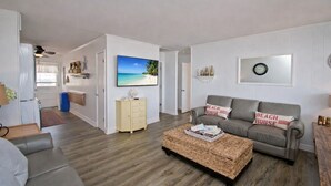 A living room with a sofa, a coffee table, a TV, and a painting of a beach scene. - At Anchor East, Oceanfront Duplex in Emerald Isle, NC! (Emerald Isle)