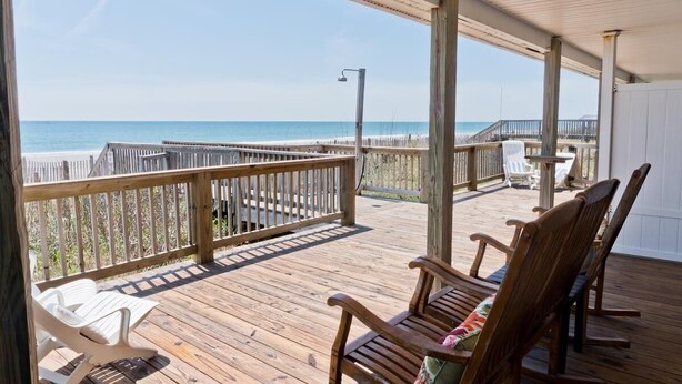 A wooden deck with a view of the beach, wooden chairs, and a white table. - At Anchor East, Oceanfront Duplex in Emerald Isle, NC! (Emerald Isle)
