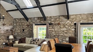 A stone-walled room with a high ceiling, a sofa, a wooden table, and a window with a view of greenery.