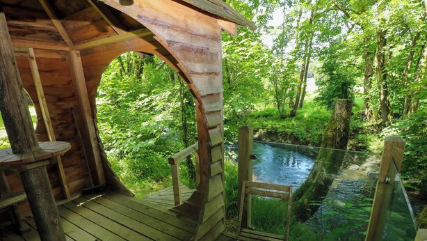 A wooden deck with a glass railing overlooking a river, surrounded by lush greenery.