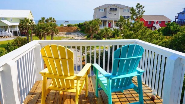 Une terrasse en bois avec deux fauteuils Adirondack, une balustrade blanche et une vue sur les maisons et l’océan.
