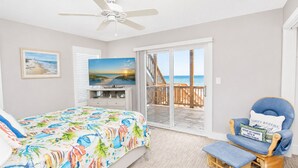 A bedroom with a bed, a TV, a chair, and a balcony with a view of the beach. - Atlantic Sea Oats, Oceanfront Duplex in Emerald Isle, NC! (Emerald Isle)