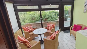 A sunlit patio with wicker furniture, a tiled floor, and a glass door leading to a garden.