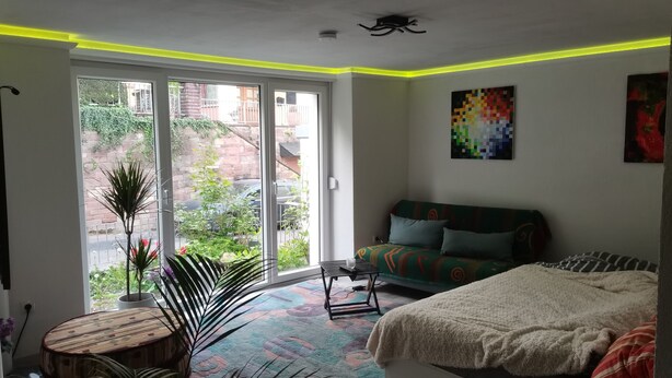 A modern bedroom with a large sliding glass door, a colorful sofa, a small round table, and a cozy bed.