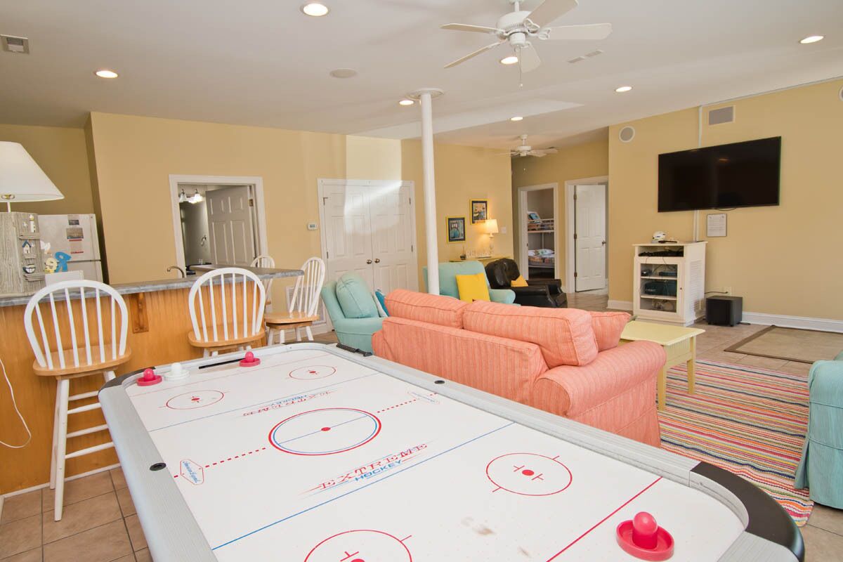 A recreational room with a shuffleboard table, seating area with chairs, a sofa, and a television mounted on the wall.