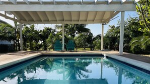 A clear swimming pool with a wooden deck and a white pergola overhead.
