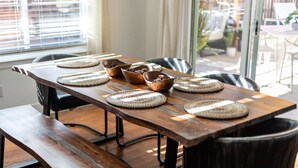 Une table à manger en bois, agrémentée de sets de table et de bols en fibres tressées, placée dans une pièce dotée de grandes fenêtres et offrant une vue sur un espace de détente extérieur.