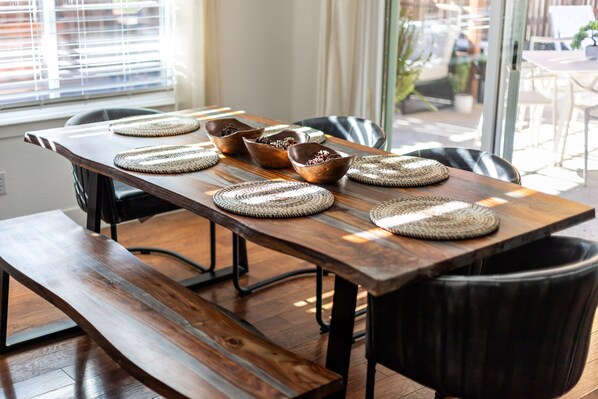 A wooden dining table with woven placemats and bowls, set in a room with large windows and a view of outdoor seating.
