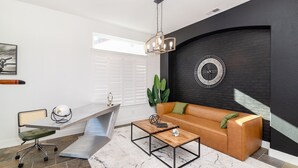 Un salon moderne avec un mur en briques noires, une horloge ronde, un canapé en cuir marron, une table basse et un bureau avec une chaise.