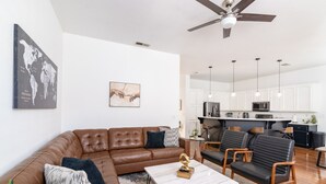 Un salon moderne avec un canapé d’angle en cuir marron, une table basse en marbre et deux fauteuils en cuir noir. On peut voir un ventilateur de plafond et des lampes suspendues.