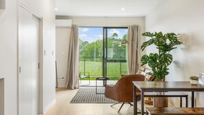 A modern room with a dining table, a brown chair, a large potted plant, and a view of a green landscape through a sliding glass door. - Urban Pulse Townhouse with parking (Auckland South)