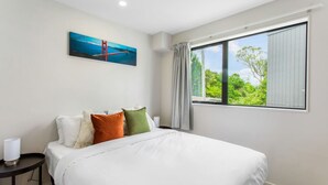 A hotel room with a large bed, a painting of a bridge, a window with curtains, and a nightstand with a lamp. - Urban Pulse Townhouse with parking (Auckland South)
