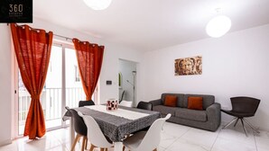 Un salon moderne avec un canapé gris, des coussins rouges, une table à manger avec des chaises et une grande fenêtre ornée de rideaux orange.