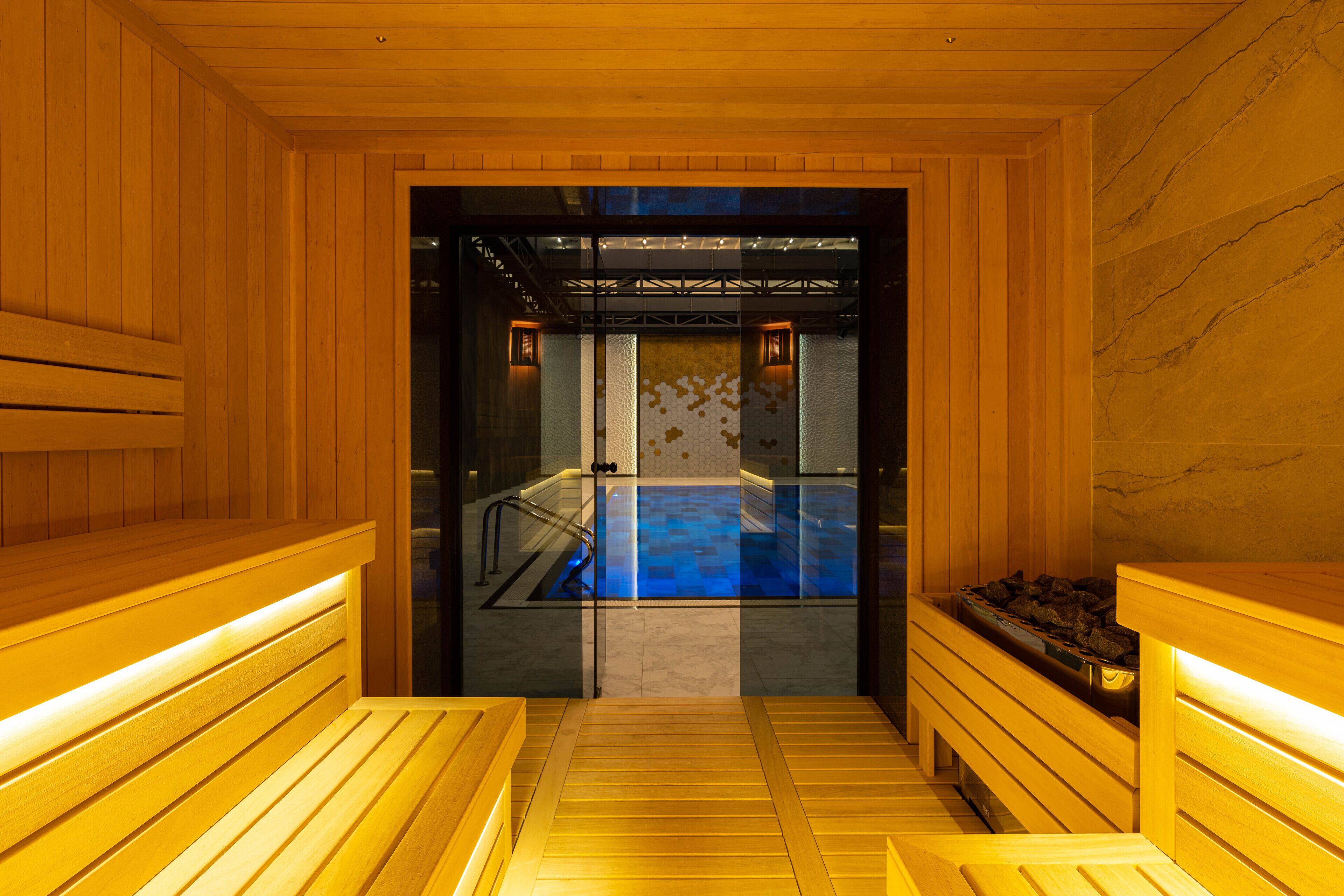 A sauna with wooden benches and a glass door leading to a blue pool area.