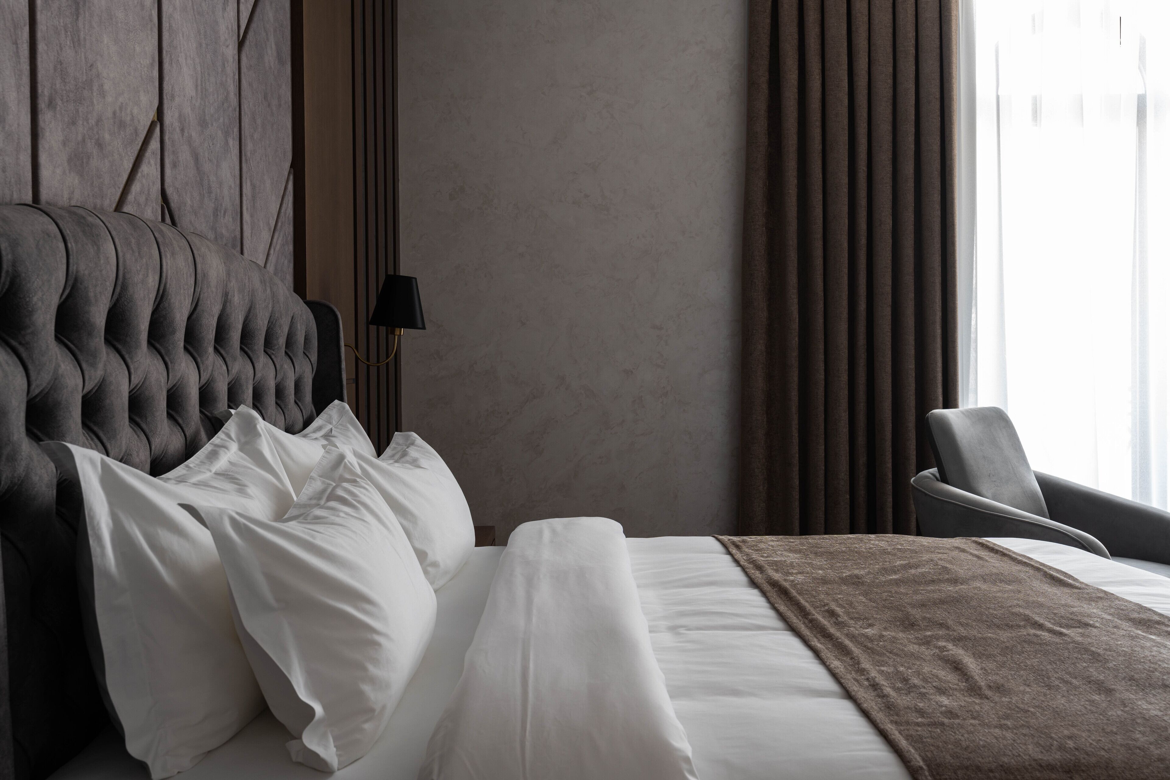 A neatly made bed with white linens and a brown throw blanket, a modern upholstered headboard, a side table with a lamp, and a view of the outdoors through sheer curtains.