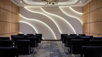 Sala de conferência com fileiras de cadeiras pretas, carpete estampado e design moderno de teto com iluminação embutida.