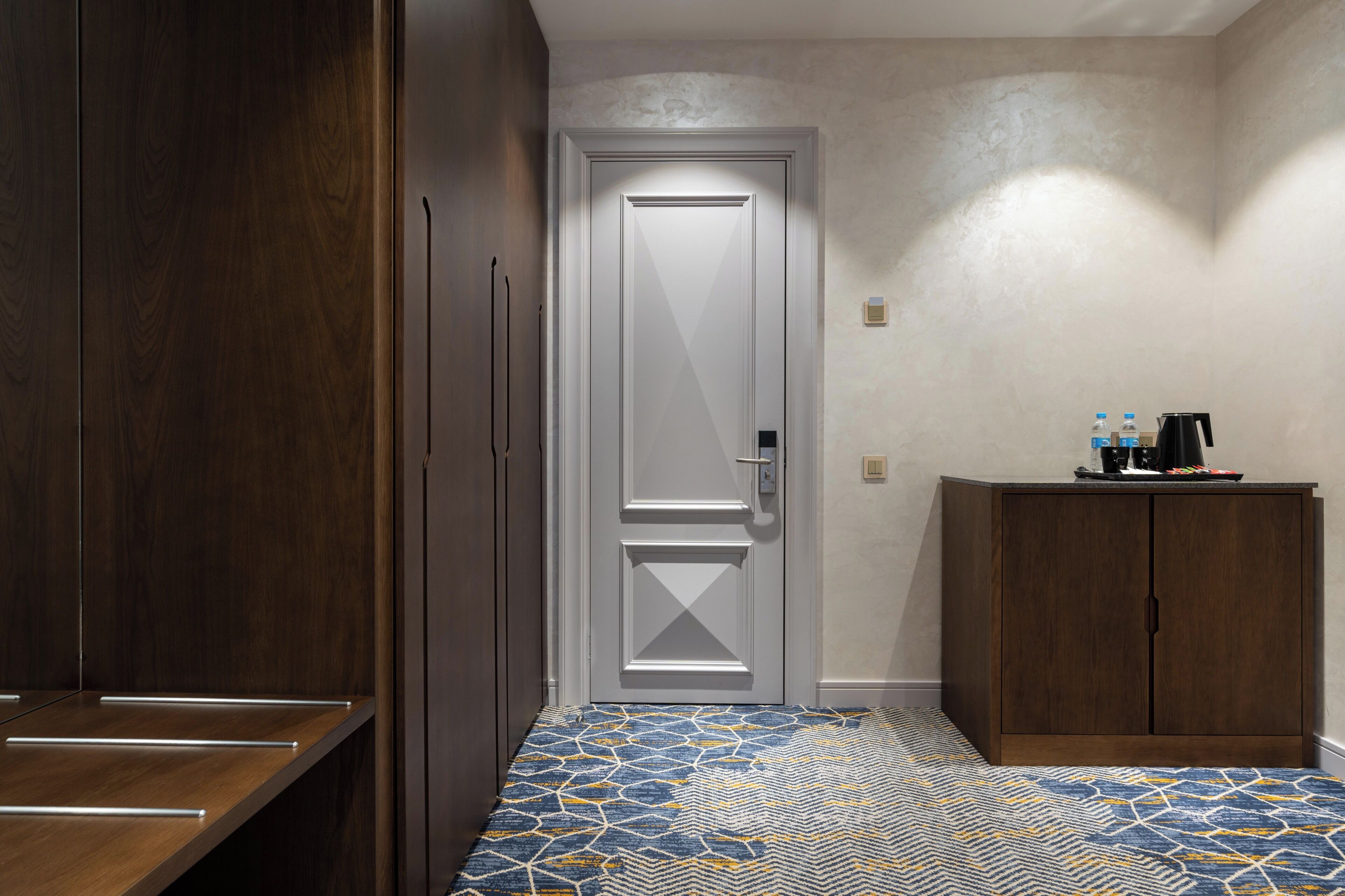 A hallway with a patterned floor, a closed door, a cabinet with a coffee maker, and a wooden wardrobe.