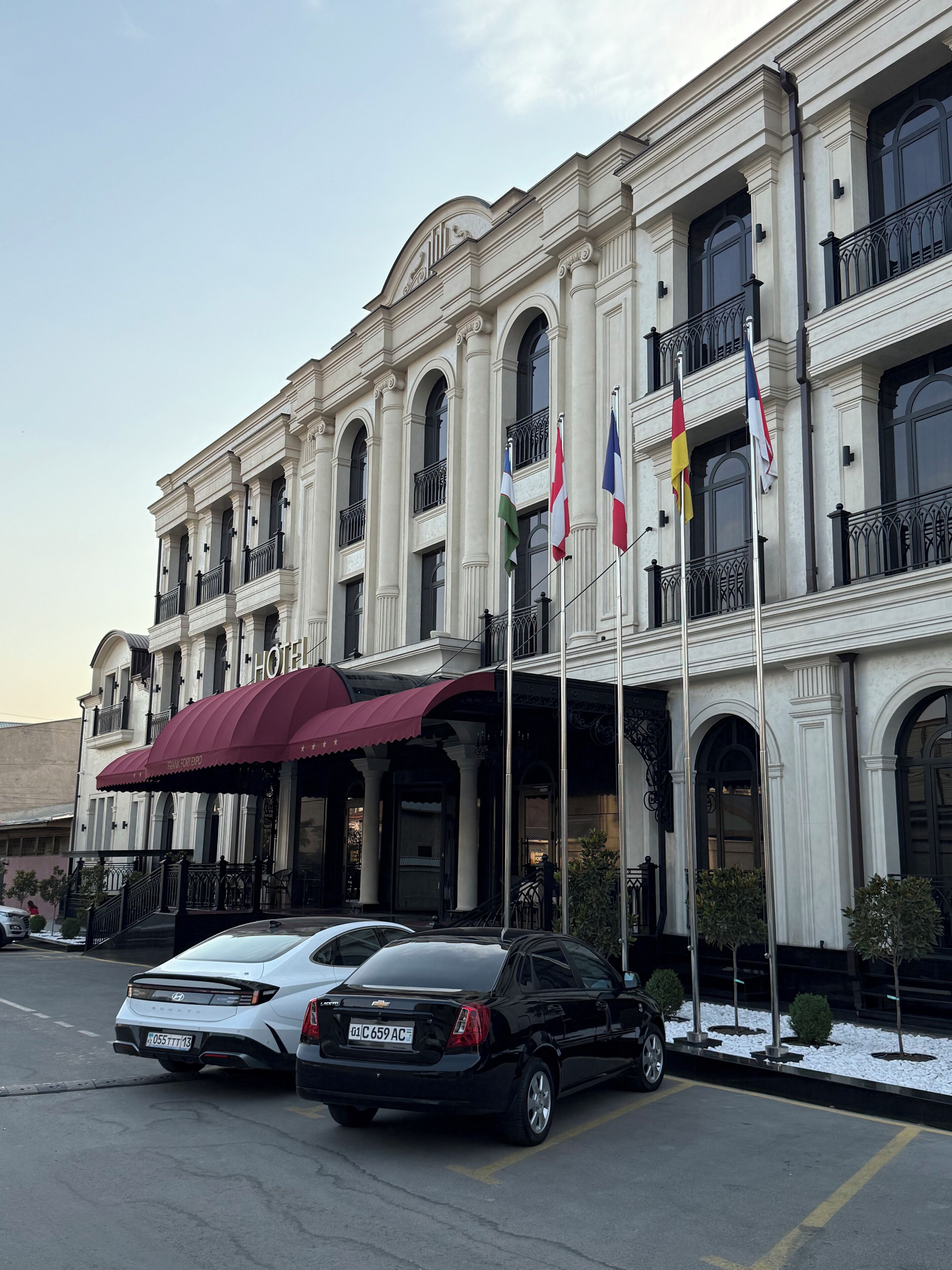 A hotel with a classic facade, multiple balconies, and flags. There are cars parked in front.