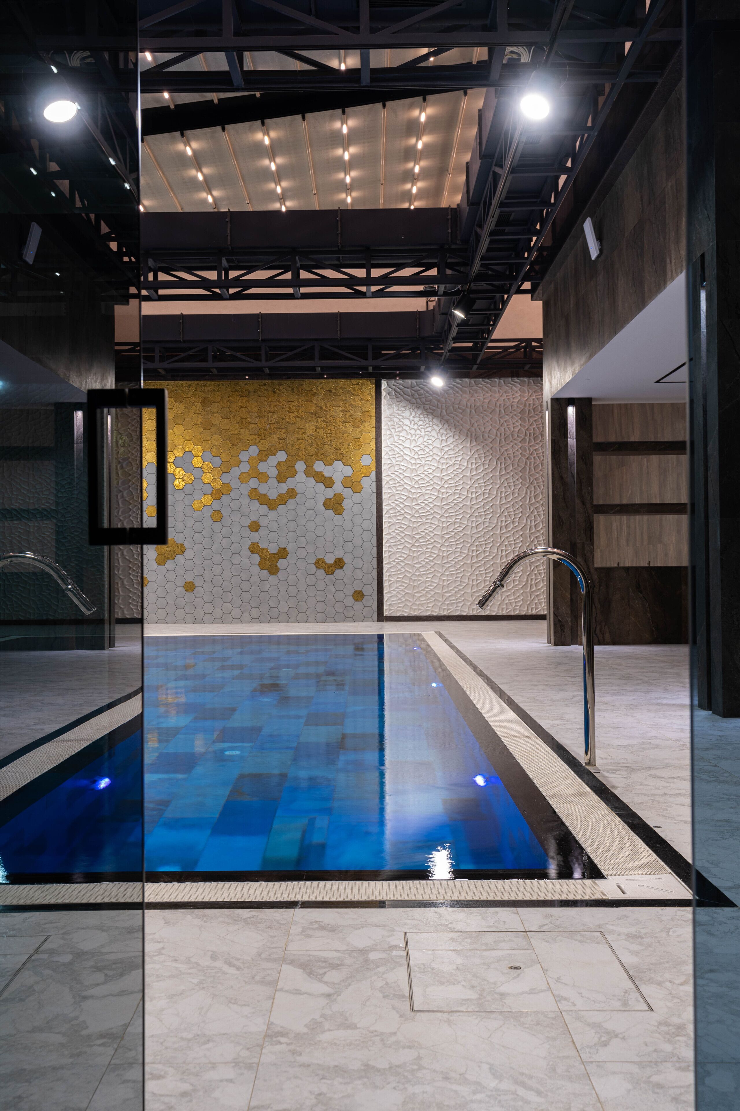 A modern indoor swimming pool with a geometric tile pattern, a wall with hexagonal gold accents, and a metal faucet.