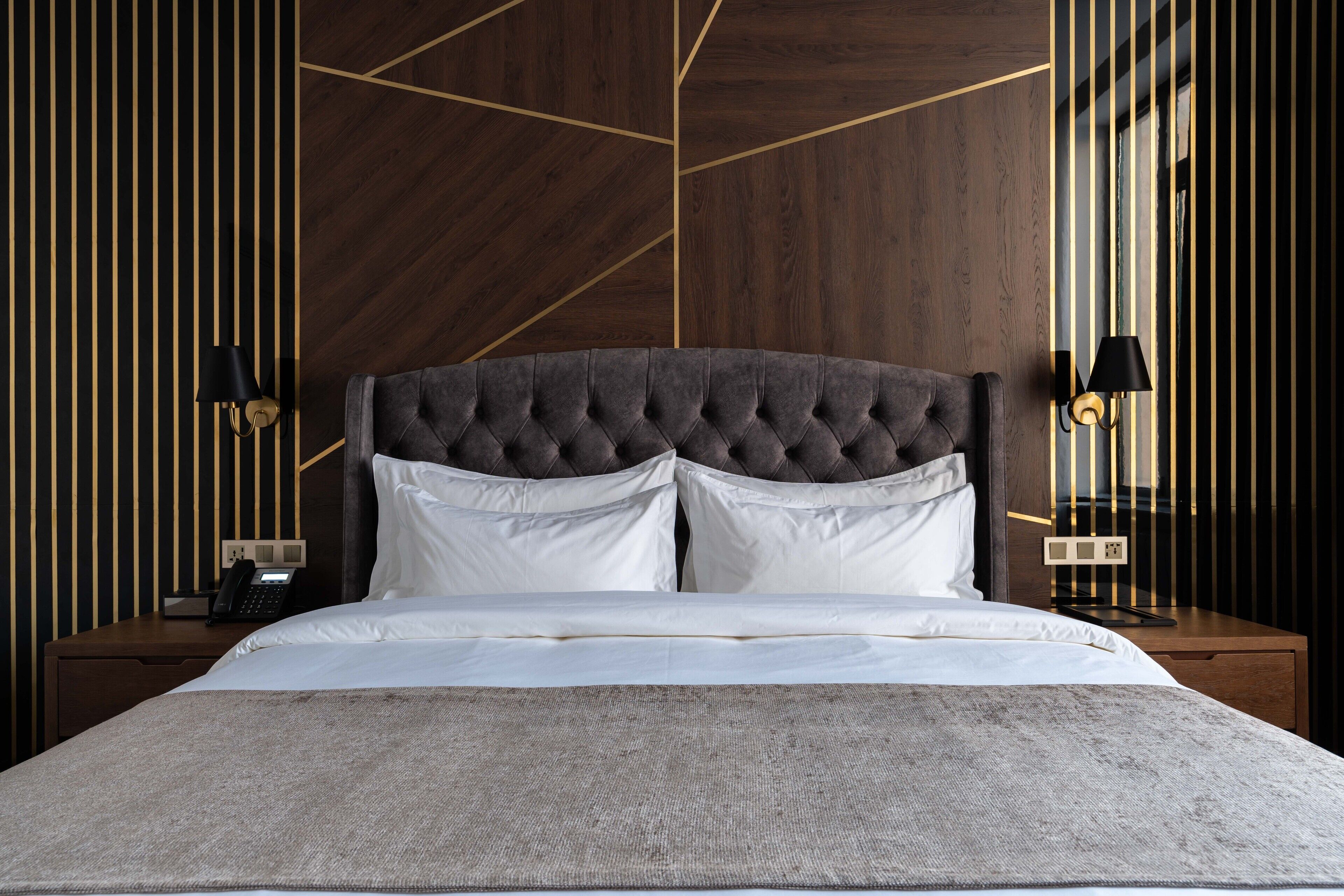 Een netjes opgemaakt bed met een gestuufd hoofdbord, witte kussens en een grijze deken. Er staan twee nachtkastjes met zwarte lampen en een telefoon op één ervan. De muur heeft een geometrisch patroon met donkerhouten panelen.